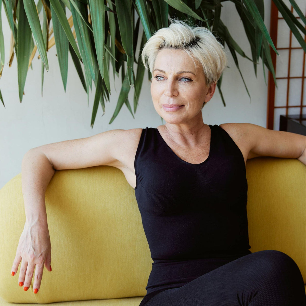 Perimenopausal Woman in black outfit sitting on a yellow couch with green plants in the background
